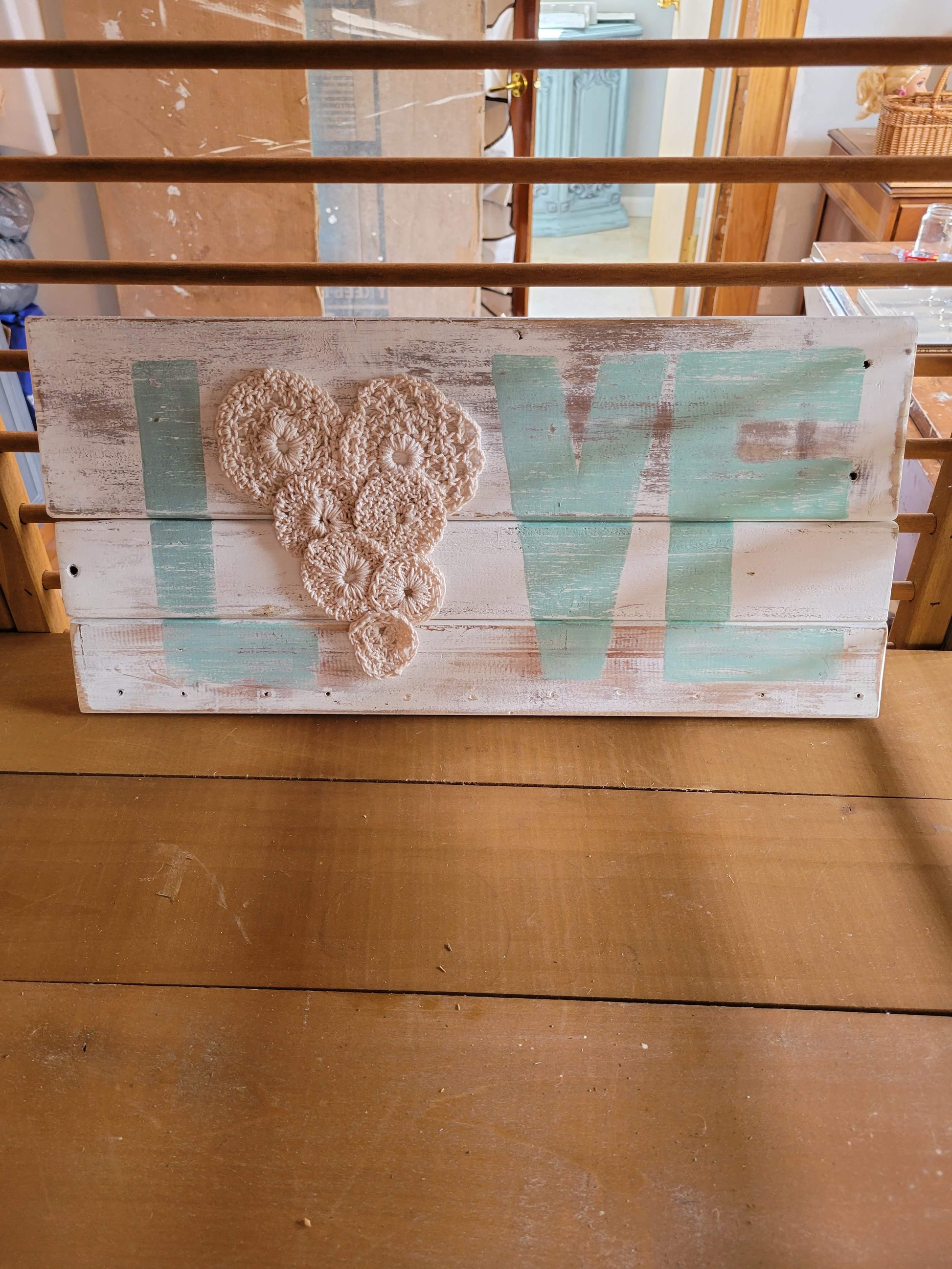 White washed pallet wood sign, "Love," painted light blue letters. The O is replaced by small crocheted flowers arranged in a heart shape.