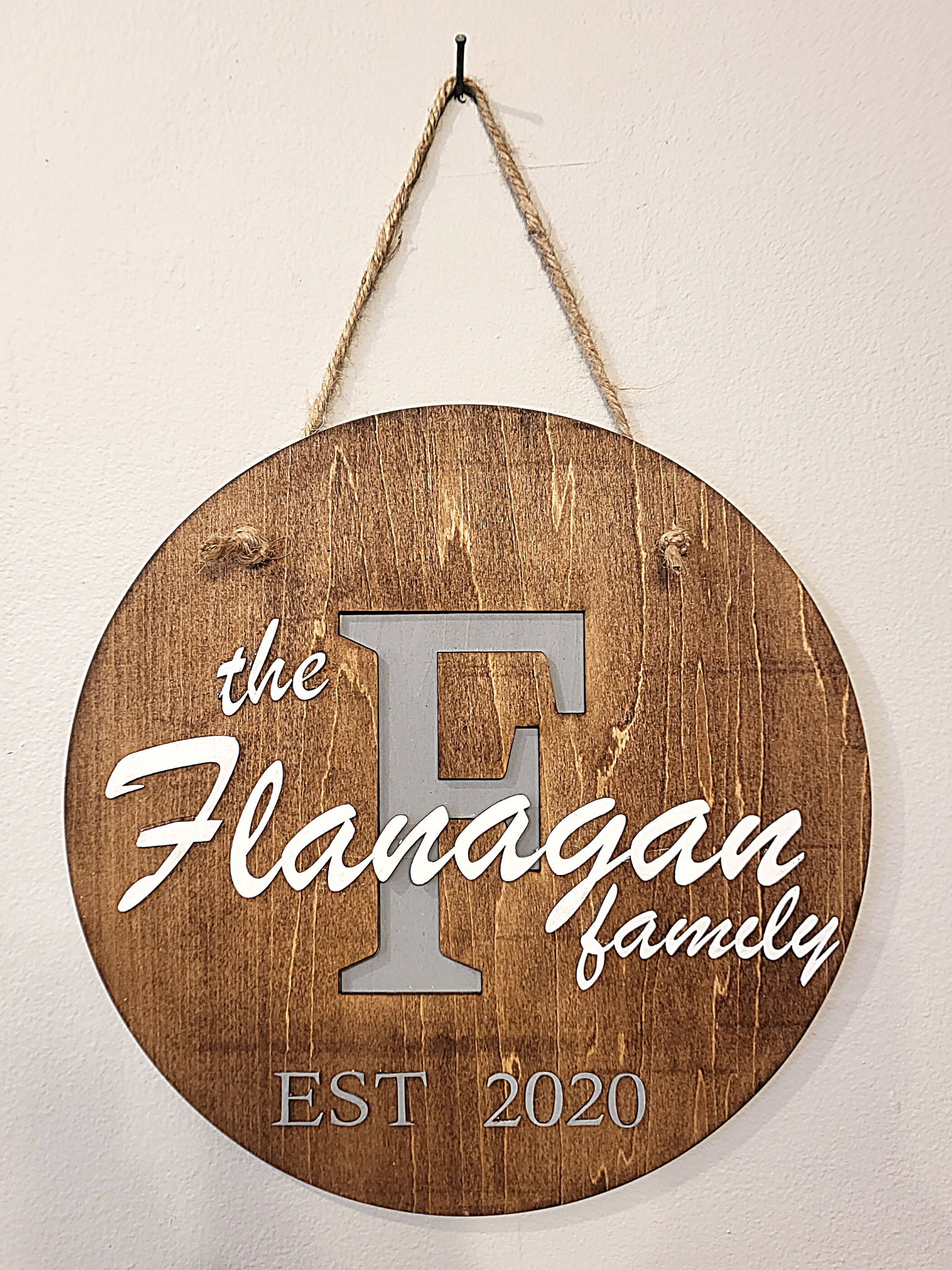 Personalized round wooden name sign. Customize with your family name and date established. Background of letter is painted light gray and laser cut name is painted white. Approximately 11" sign. dark stained wood, natural jute rope hanger.