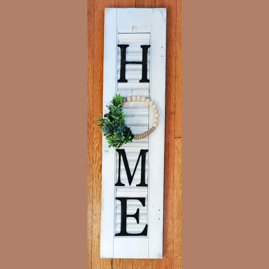 Small white shutter that says "HOME" with a small beaded wreath in place of the O.