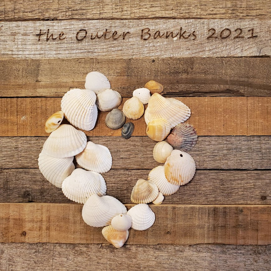 Seashells gathered from the beach positioned in the shape of a heart on laser engraved pallet wood that reads the outer banks 2021