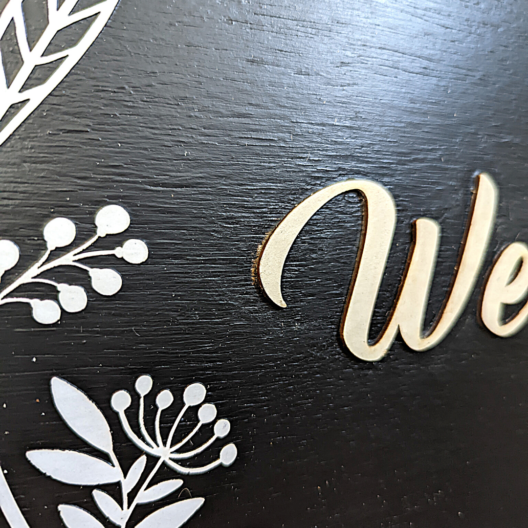 This black wooden Welcome sign has bohemian vibes with the three dimensional paper flower detail and multi-colored wooden beads. The "Welcome" is laser cut wood.